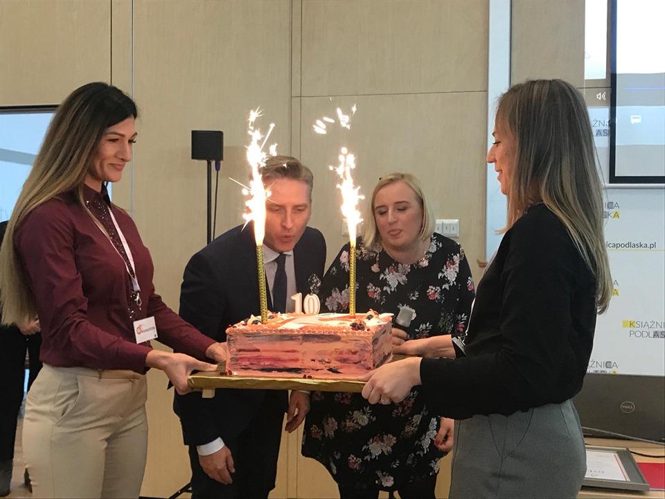Dwie kobiety trzymają tort urodzinowy z racami. zdmuchnięcie świeczek na urodzinowym torcie przez i Dyrektorkę Oddziału Magdalenę Bobrowską oraz  Prezesa Zarządu Przemysława Żydoka