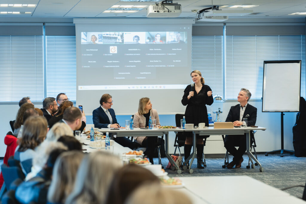 Zdjęcie przedstawia formalne spotkanie biznesowe lub konferencję odbywającą się w nowoczesnej sali konferencyjnej. Uczestnicy siedzą przy długim stole, na którym znajdują się butelki wody, filiżanki, dokumenty oraz talerze z przekąskami. W tle widać ekran projekcyjny z wyświetlonym spotkaniem online, na którym widoczne są nazwiska i zdjęcia uczestników zdalnych. Kobieta w czarnej sukience stoi i przemawia do zgromadzonych, trzymając mikrofon. Obok niej siedzi mężczyzna w eleganckim garniturze, który uważnie słucha. Pozostali uczestnicy, ubrani w formalne stroje, również słuchają lub notują. W tle znajdują się okna z zamkniętymi żaluzjami oraz flipchart na stojaku