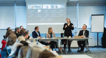 Zdjęcie przedstawia formalne spotkanie biznesowe lub konferencję odbywającą się w nowoczesnej sali konferencyjnej. Uczestnicy siedzą przy długim stole, na którym znajdują się butelki wody, filiżanki, dokumenty oraz talerze z przekąskami. W tle widać ekran projekcyjny z wyświetlonym spotkaniem online, na którym widoczne są nazwiska i zdjęcia uczestników zdalnych. Kobieta w czarnej sukience stoi i przemawia do zgromadzonych, trzymając mikrofon. Obok niej siedzi mężczyzna w eleganckim garniturze, który uważnie słucha. Pozostali uczestnicy, ubrani w formalne stroje, również słuchają lub notują. W tle znajdują się okna z zamkniętymi żaluzjami oraz flipchart na stojaku