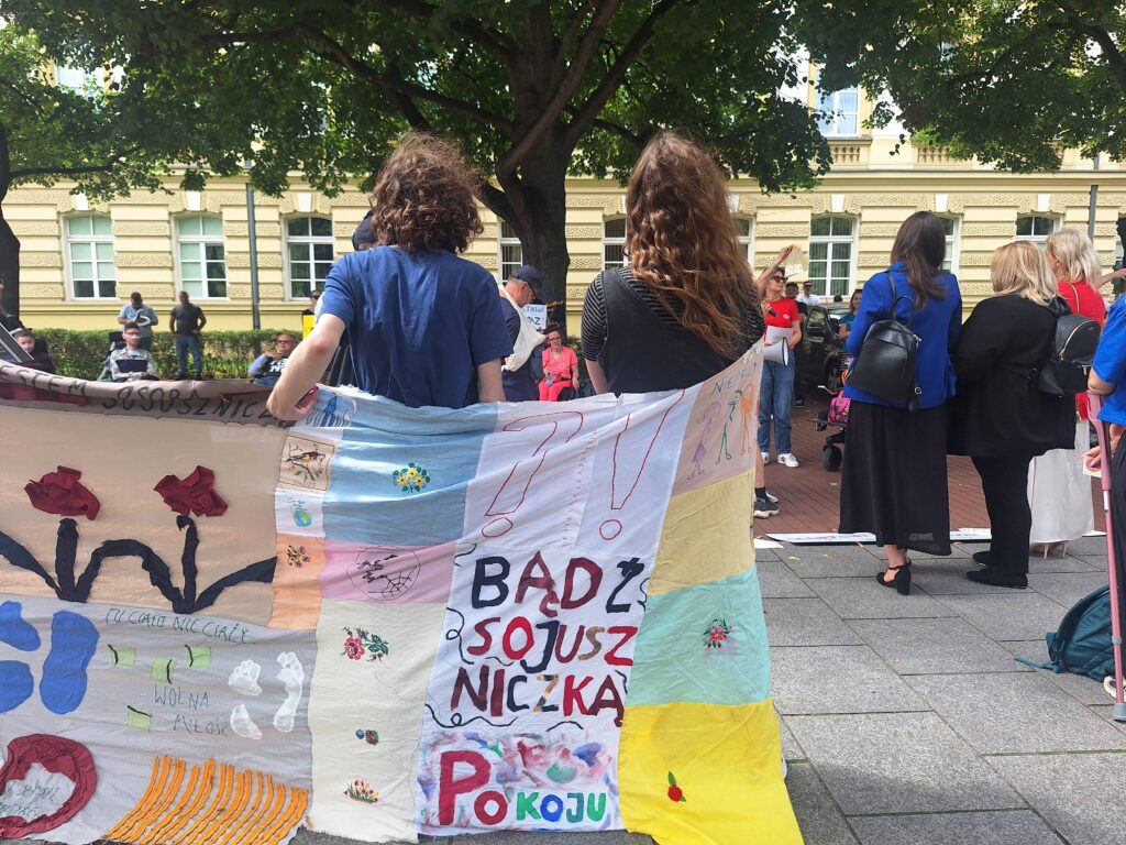 Dwie osoby trzymają kolorowy transparent z napisem „Bądź sojuszniczką pokoju” podczas demonstracji pod drzewami, wokół zgromadzeni ludzie.