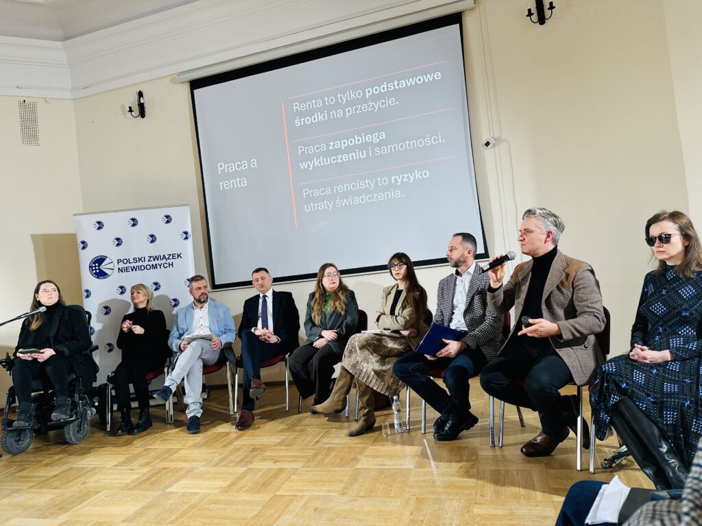 Zdjęcie z Konferencji prasowej. Osob wypowiadajace się siedzą na krzesłach nad ich głowami ekran z prezentacją. Mikrofon trzyma Przemysław Żydok.