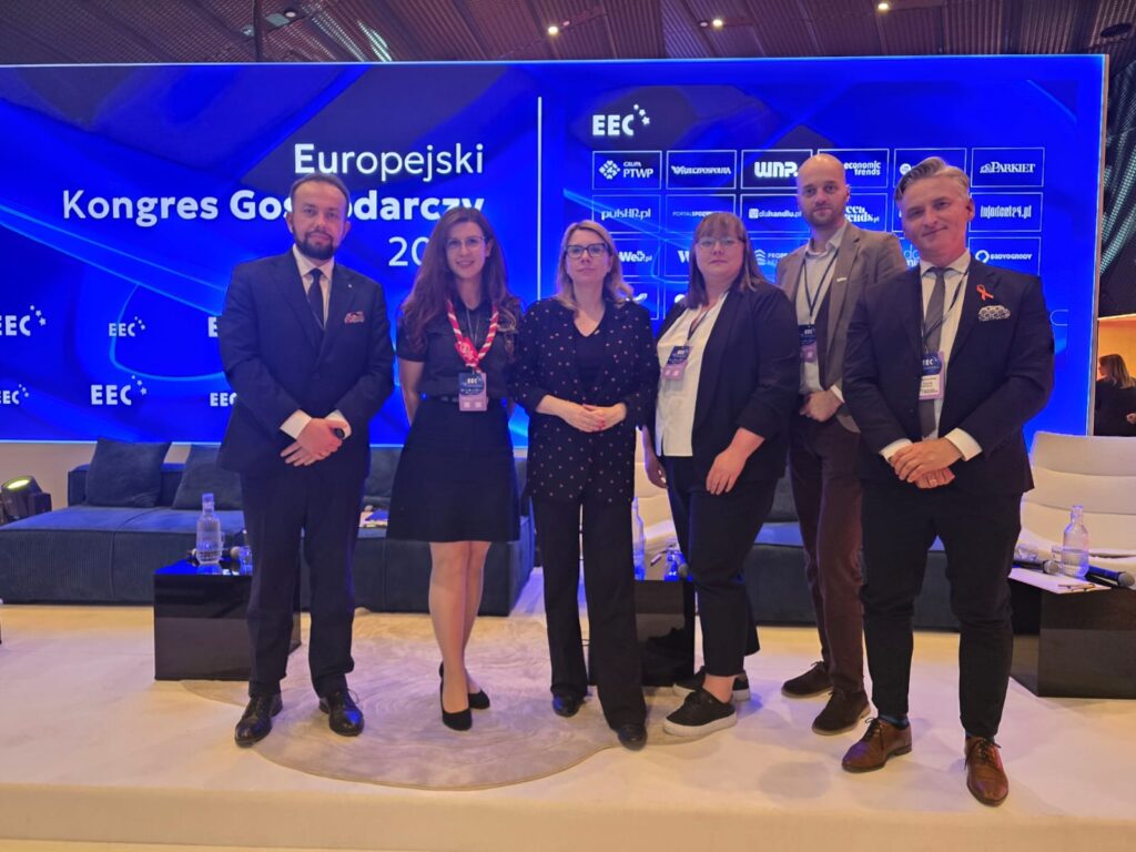 Grupa sześciu osób w formalnych strojach stoi na scenie podczas wydarzenia „Europejski Kongres Gospodarczy (EEC)”. W tle widoczny jest duży, niebieski ekran z logo wydarzenia i partnerów. Uczestnicy mają identyfikatory konferencyjne i pozują do wspólnego zdjęcia.