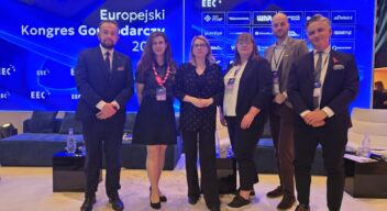 Grupa sześciu osób w formalnych strojach stoi na scenie podczas wydarzenia „Europejski Kongres Gospodarczy (EEC)”. W tle widoczny jest duży, niebieski ekran z logo wydarzenia i partnerów. Uczestnicy mają identyfikatory konferencyjne i pozują do wspólnego zdjęcia.