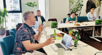 Mężczyzna prezentuje zagadnienie przy stole konferencyjnym, gestykulując podczas wypowiedzi. Pozostali uczestnicy słuchają i pracują na laptopach.