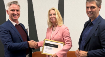 Trzy osoby stoją przed pomarańczową sofą na tle ściany z czarno-białym wzorem. Dwie osoby w centrum ściskają sobie dłonie na znak zawarcia partnerstwa, a jedna z nich trzyma oprawiony dokument. Wszystkie osoby uśmiechają się i są ubrane w stroje biznesowe.